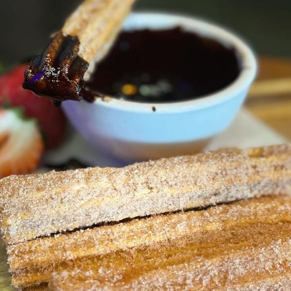 A churro dipped in chocolate sauce, with more churros and strawberries in the foreground.